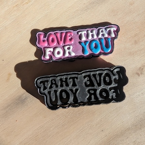 May include: Two layered pins with the text "LOVE THAT FOR YOU". The top pin features pink, white, and blue lettering with a black outline. The bottom pin is a black outline of the same design.