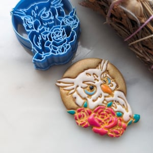 May include: A blue plastic cookie cutter shaped like an owl with roses around it. A white owl cookie with pink roses is also shown.