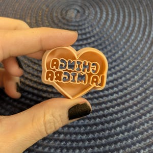 May include: A heart-shaped cookie cutter in a light peach color. The text "LA CHINCU" is embossed in a retro font with orange and blue accents. The cookie cutter is held against a blue textured background.
