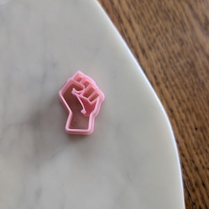May include: A small, pink fist-shaped cookie cutter. The cutter is made of plastic and sits on a white marble surface. The background is a wooden table.