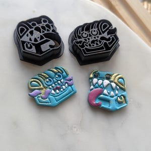 May include: Four pieces: two black plastic cutters and two blue, gold, and purple monster-shaped clay figures. The cutters have a detailed monster face design. The clay figures have monster faces with tongues and teeth.