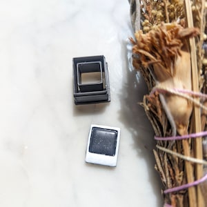 May include: Two small, black square-shaped objects, one nested inside the other, sit next to a small, white-framed square with a black center. A bundle of dried herbs with a pink and purple string is in the background.