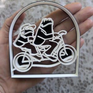 Puede incluir: Un cortador de galletas de plástico blanco con un diseño de relieve de dos figuras montando una bicicleta tándem. El diseño está encerrado dentro de un marco de arco redondeado, con las figuras y la bicicleta representadas en un estilo lineal.