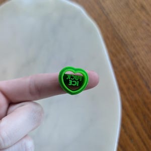 Puede incluir: Un cortador de galletas verde en forma de corazón con las palabras "ICE LUCK" en verde, sostenido por un dedo. El cortador está hecho de plástico y tiene un borde liso y redondeado. El fondo es una superficie de color claro y una mesa de madera.