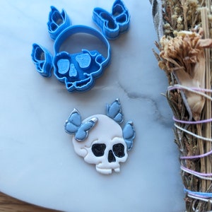 May include: Two blue plastic skull-shaped cookie cutters with butterfly wings, and a white skull with blue butterfly wings. The skull has black eye sockets. The items are on a light gray surface with a bundle of dried herbs.