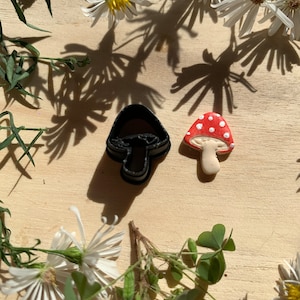 May include: A black mushroom cookie cutter and a red and white mushroom cookie on a wooden surface with white flowers and green leaves.