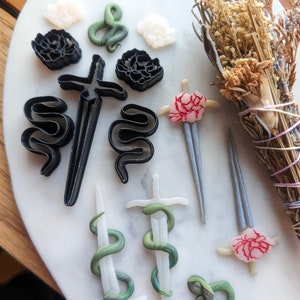 May include: A collection of black, white, and green polymer clay charms featuring snakes, swords, and flowers. The charms are arranged on a white marble surface.