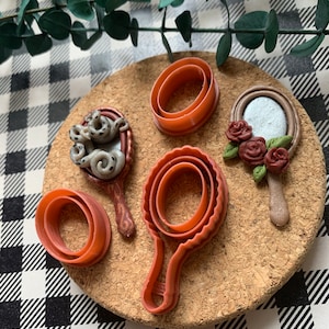 May include: Four red plastic cookie cutters shaped like hand mirrors. One mirror has a white and gray octopus design, another has a red rose design, and the other two are plain.