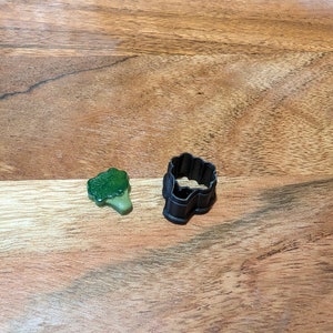 May include: A green plastic broccoli cookie cutter and a green plastic broccoli sweet on a wooden surface.