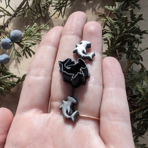 May include: Three silver hammerhead shark charms, two are flat and one is a 3D cutout. The charms are in a hand.