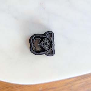 May include: A black plastic cookie cutter in the shape of a stylized animal face, featuring large eyes and ears. The cutter is placed on a white surface, likely for baking or crafting.