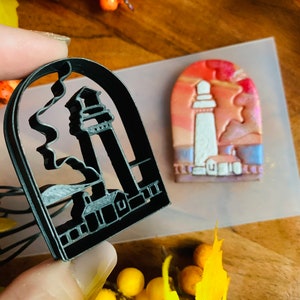 May include: A black plastic cookie cutter shaped like a lighthouse with a wavy line representing the ocean in the background. The cutter is held in a hand, and a small, painted lighthouse is visible in the background.