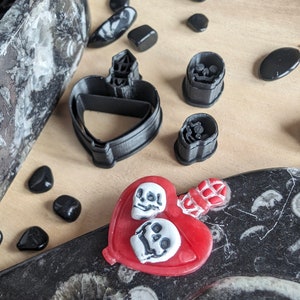 May include: A collection of Halloween-themed craft supplies. Includes black plastic heart-shaped cookie cutters, skull-shaped molds, and a red heart-shaped pendant with white skull designs. Black stones and a decorative container are also present.