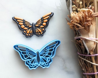 May include: Two butterfly cookie cutters, one black and orange and one blue. The cutters are on a white marble surface.