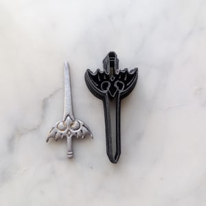 Puede incluir: Dos cortadores de galletas de espada de color plateado, uno más grande que el otro, sobre una superficie de mármol blanco.