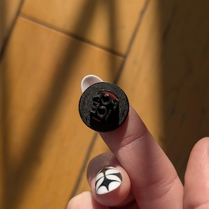 May include: A small, round, black object with the word "LOVE" in red and black lettering. The object is held between two fingers, with a background of wooden flooring.