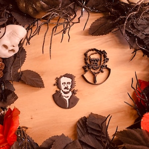 Puede incluir: Dos cortadores de galletas negros con temática de Edgar Allan Poe sobre una superficie de madera, rodeados de hojas negras, rosas rojas y decoraciones de Halloween. Un cortador presenta el retrato de Poe, el otro un busto con un marco floral.
