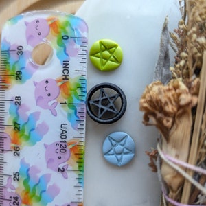 May include: Three small pentagram charms, one green, one black, and one blue, are arranged on a white surface next to a ruler.