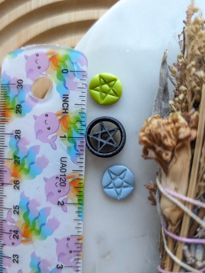 May include: Three small pentagram charms, one green, one black, and one blue, are arranged on a white surface next to a ruler.
