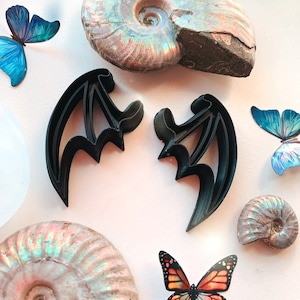 May include: Two black bat-shaped cookie cutters on a white background with iridescent ammonite fossils and blue and orange butterflies.