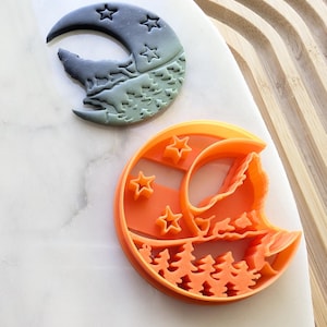 Puede incluir: Dos cortadores de galletas en forma de media luna, uno verde y otro naranja, con un lobo aullando a la luna y una escena de bosque. Ambos cortadores tienen estrellas.