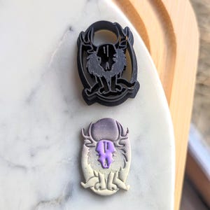 Puede incluir: Dos cortadores de galletas, uno negro y otro blanco con acentos morados, con forma de criatura con cuernos y una cara de calavera.