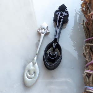 Puede incluir: Dos cucharas de plata con un diseño de luna creciente. Una cuchara es negra y la otra es plateada.