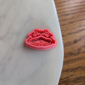 Könnte beinhalten: Ein rosa Kunststoff-Ausstecher in Form eines Froschgesichts. Der Ausstecher hat detaillierte Augen, einen Mund und strukturierte Kanten. Er liegt auf einer weißen Marmoroberfläche.