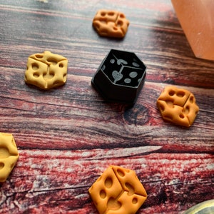 May include: A collection of cheese-shaped polymer clay charms in various shades of yellow and orange. One black, geometric mold is also present. The charms are scattered on a wooden surface, with a pink crystal and a dark stone in the background.