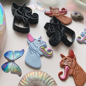 May include: Five ceramic cat head sculptures with open mouths and long tongues. The sculptures are in various colors, including black, brown, blue, and pink. The tongues are painted with a green and pink swirl pattern.