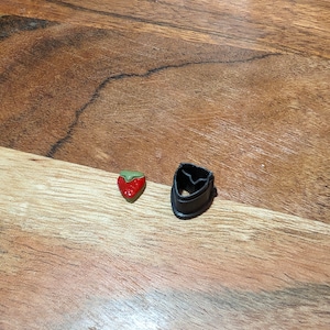 May include: Two cookie cutters, one shaped like a heart and the other shaped like a strawberry. The heart cutter is black and the strawberry cutter is red, green, and yellow.