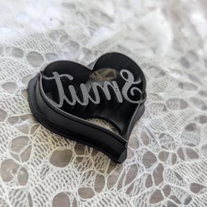 Peut inclure: Un emporte-pièce en forme de cœur noir avec le mot "June" écrit en lettres cursives intégré au centre. L'ustensile possède un bord surélevé pour la découpe et une section intérieure en relief pour imprimer le texte dans la pâte ou l'argile.