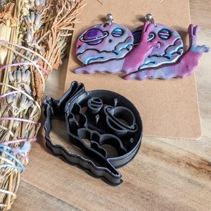 May include: A pair of polymer clay earrings shaped like snails with a galaxy design. The snails are painted in shades of purple, pink, and blue with silver accents. The earrings are hanging from a brown card with a hole punched in the top.