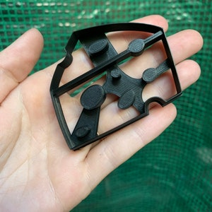 May include: A black plastic cookie cutter in the shape of a wedge of cheese, held in a hand. The cutter has several oval and circular cutouts, and a rectangular shape. The background is a green mesh.
