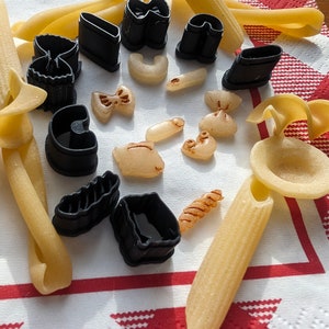 May include: A collection of black plastic pasta cutters and various shapes of dry pasta on a red and white checkered tablecloth.