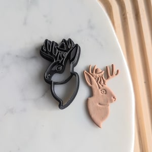 Puede incluir: Un cortador de galletas de ciervo negro y marrón y una galleta de ciervo marrón. El cortador de galletas está hecho de plástico y tiene un diseño detallado. La galleta está hecha de masa y tiene un diseño simple.