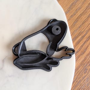 May include: A black plastic frog-shaped cookie cutter. The cutter features the frog's eyes, mouth, and webbed feet. The frog is lying down on a white surface.