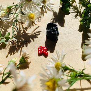 May include: A small black mushroom-shaped cookie cutter and a red and white mushroom-shaped cookie on a wooden surface with white flowers.