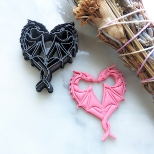 Puede incluir: Dos figuras de dragón en forma de corazón, una negra y otra rosa, con detalles intrincados. Los dragones se miran con la cabeza tocando.