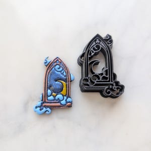May include: Two 3D printed cookie cutters in the shape of a gothic arch window with a crescent moon and clouds inside. The cutter on the left is painted with blue and gold accents.
