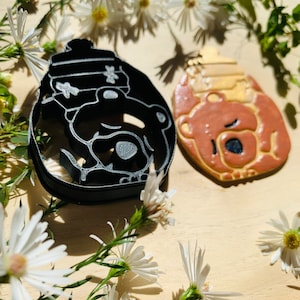 May include: Two bear-shaped cookie cutters, one black and one brown, are on a wooden surface. The black cutter is a silhouette, while the brown one is filled in with details. White flowers and green foliage surround the cutters.
