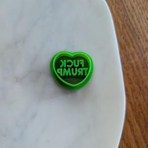 May include: A green heart-shaped cookie cutter with the words "FUCK TRUMP" in green, reversed text. The cutter is on a white marble surface.
