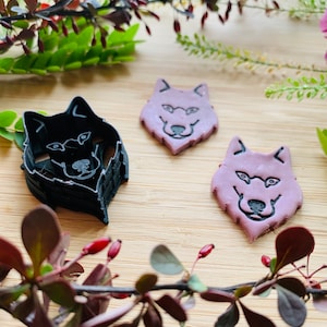 May include: Two purple wolf-shaped cookie cutters and one black wolf-shaped cookie cutter on a wooden surface. The cookie cutters are surrounded by green and red leaves.