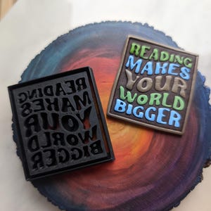 Puede incluir: Dos letreros rectangulares negros y marrones con el texto "Reading makes your world bigger" en diferentes colores y fuentes. Los letreros están sobre una superficie de madera colorida.