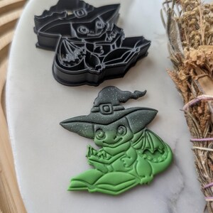 Puede incluir: Dos cortadores de galletas con forma de dragón con un sombrero de bruja. El cortador superior es negro y el inferior es verde.