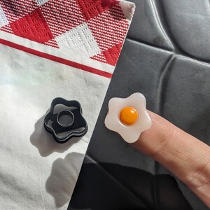 May include: A white plastic ring shaped like a fried egg with a yellow yolk in the centre. A black plastic ring shaped like a flower is also in the image.