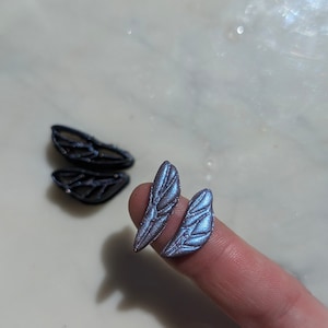 Puede incluir: Dos alas de hada iridiscentes de color azul y morado con una superficie texturizada. Las alas son pequeñas y delicadas, y son perfectas para agregar un toque de magia a cualquier proyecto artesanal.
