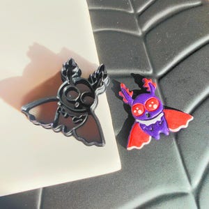 May include: Two black plastic cookie cutters shaped like moths. One cutter is a simple outline of a moth, while the other is a more detailed design with red and purple accents.