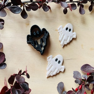 Puede incluir: Tres cortadores de galletas de fantasmas de Halloween, dos blancos y uno negro, sobre una superficie de madera con una rama de plantas de hojas rojas.