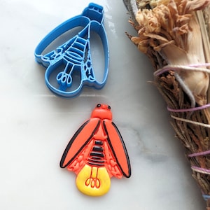 Puede incluir: Un cortador de galletas de plástico azul con forma de luciérnaga y una galleta de luciérnaga roja y amarilla.